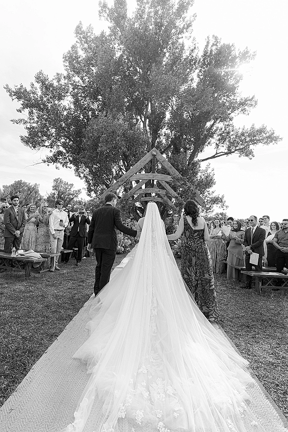 Wedding processional as bride walking down aisle with cathedral veil and long train toward a wooden floral arch on a lawn with guests standing