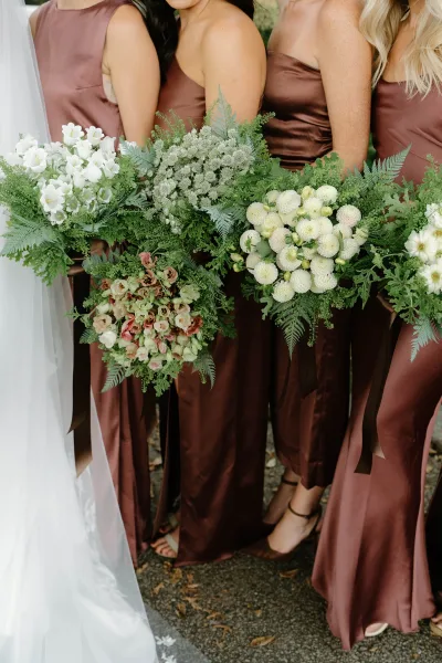 Bridesmaid bouquets with greenery and blush-white blooms held by bridesmaids in brown dresses, with ribbon streamers on leaf-strewn pavement