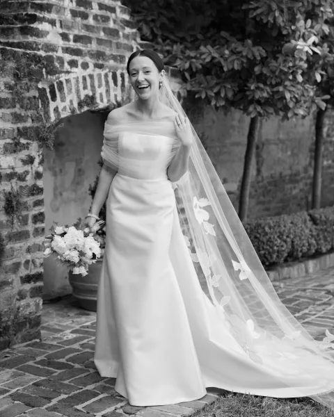 Bridal portrait of a laughing bride holding a white bouquet, wearing a strapless gown and long lace veil by a brick wall garden path