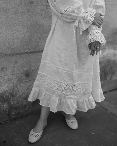 Engagement ring photo showing a hand with engagement ring in lace sleeve with ribbon ties, near woven flats by a stone wall sidewalk
