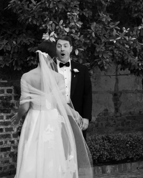 First look moment as bride in wedding dress and veil reveals herself, groom in tuxedo reacts by leafy greenery and brick wall backdrop