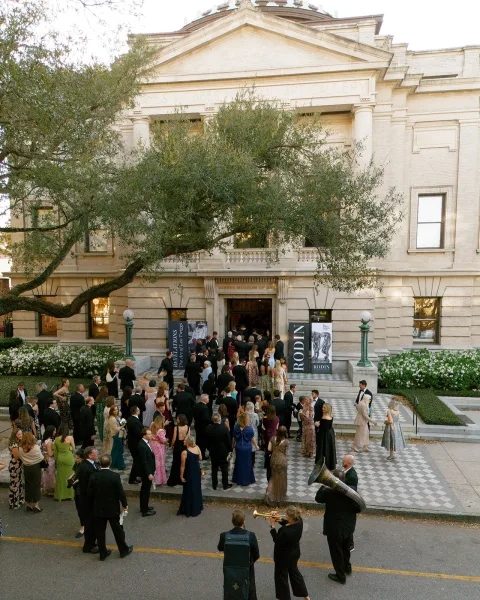 Wedding cocktail hour with wedding guests mingling in black-tie attire outside a museum entrance, brass band by checkered courtyard and lamps