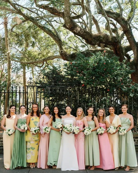 Bridesmaid group photo with pastel dresses and bouquets, posing with the bride by a wrought iron gate along a leafy garden walkway