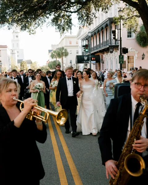 Wedding parade with bride and groom leading a second line wedding down a city street, bridal veil flowing as a brass band follows
