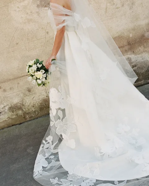 Wedding dress with off the shoulder sleeves and long floral veil, bride holding a white and green bouquet beside a stone wall sidewalk