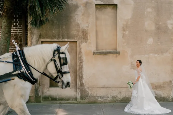 Bridal portrait of a bride with horse, laughing in an off-the-shoulder gown and veil, holding a white rose bouquet by a stucco wall