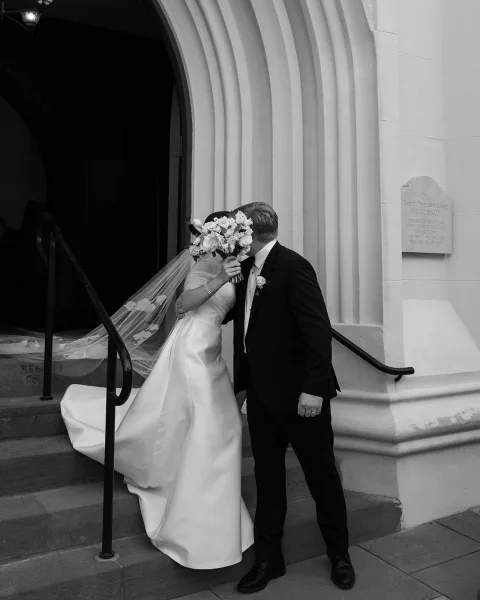 Wedding kiss as bride and groom kiss behind a white rose bouquet, her veil trailing, on stone church steps by the doorway