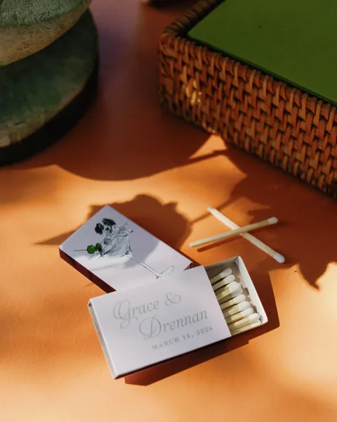 Custom matchboxes wedding matchbox favors with illustrated label and typography, set in a rattan basket on an orange tabletop with sunlit shadows