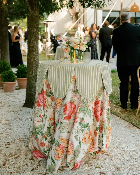 Cocktail table decor with floral centerpiece and bud vases on striped linen, surrounded by votive candles under garden string lights