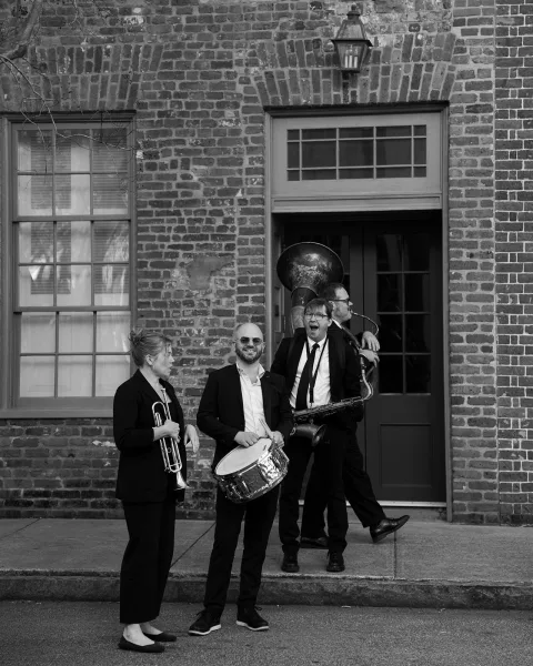 Wedding band with trumpet and saxophone posed in suits and sunglasses on a sidewalk outside a brick wall with door and window