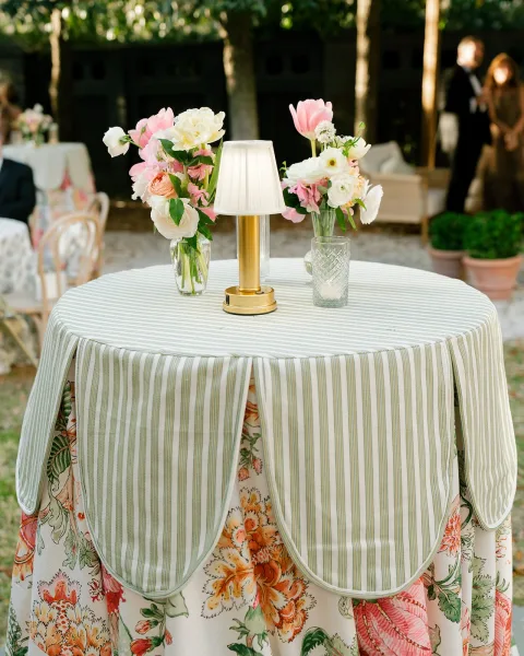 Cocktail table decor with a striped tablecloth, bud vases of pink and white flowers, and a votive candle on an outdoor patio