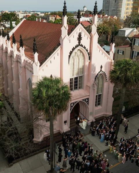 Wedding exit as newlyweds leave church steps, bride with bouquet beside groom in tux, guests cheering by iron fence on city street