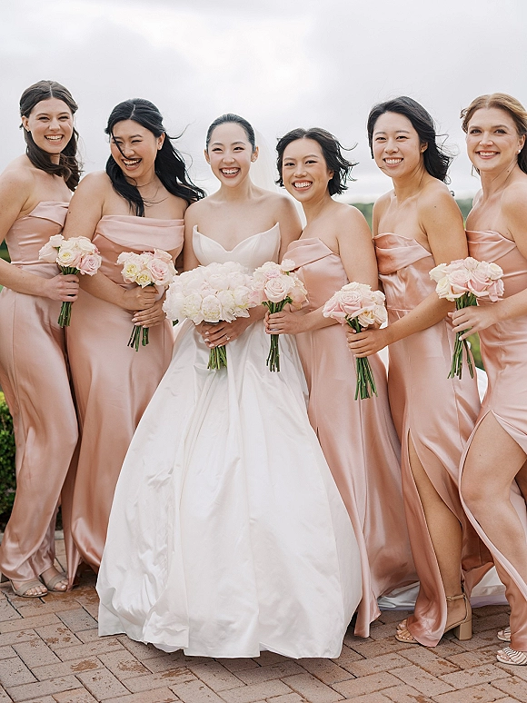 Bridesmaids group photo with bride with bridesmaids in blush satin dresses holding hydrangea and rose bouquets on a terrace with greenery