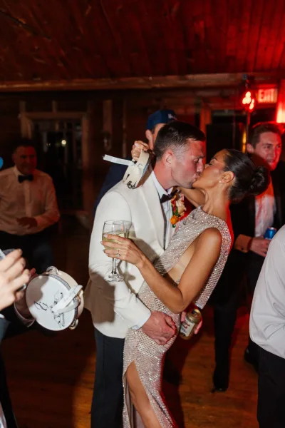 Wedding kiss as bride in a sequin beaded gown and groom in a white tuxedo share champagne on a red-lit dance floor with guests behind