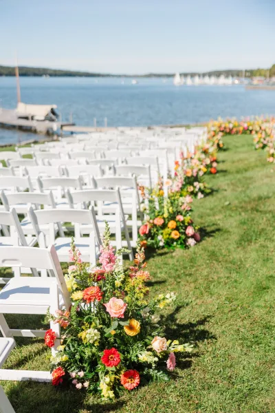 Ceremony aisle decor with white folding chair rows and flower-lined arrangements of roses and greenery on a lakeside lawn with sailboats