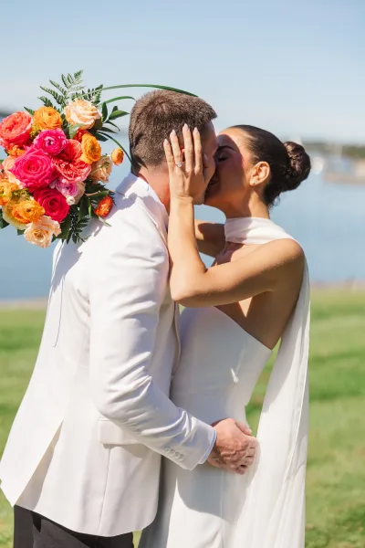 Wedding kiss portrait of bride and groom kiss by a waterfront lawn under blue sky, bride’s hand on his face showing ring and bouquet