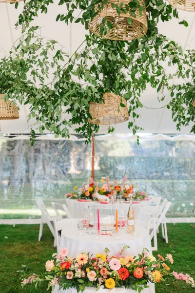 Reception tablescape in a wedding tent reception with hanging greenery, wicker pendant lights, and floral centerpieces on round white-linen tables
