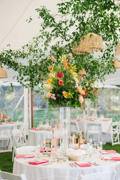 Reception tablescape with wedding table centerpiece of tall florals in a clear vase, pink napkins and glassware under wicker pendant lights in a white tent