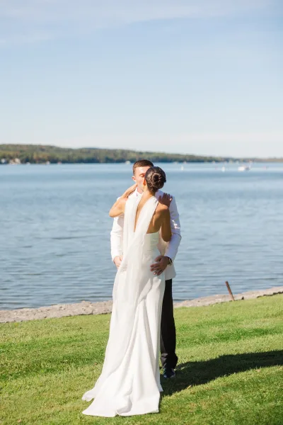 Wedding kiss portrait of bride and groom kissing on a grassy lakeshore, her veil trailing behind as he holds her waist under open sky