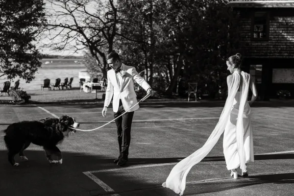 Couple with dog walking away in a black-and-white wedding portrait, bride’s long veil trailing as groom holds leash by waterfront trees