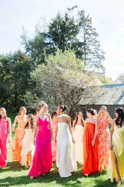 Bridesmaid group photo of bride with bridesmaids in colorful satin dresses, walking on a grass lawn with trees, blue sky, and a rustic barn behind
