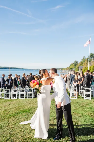 Wedding kiss as the couple embraces with a bright bouquet accent, guests cheering behind on a sunny lakeside lawn under blue sky
