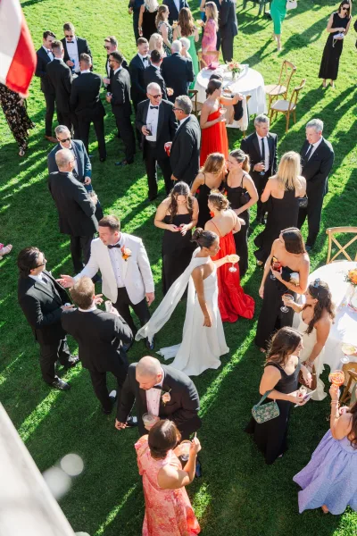 Wedding cocktail hour with guests mingling outdoors as the bride in veil moves between white tablecloth tables holding cocktails on a green lawn