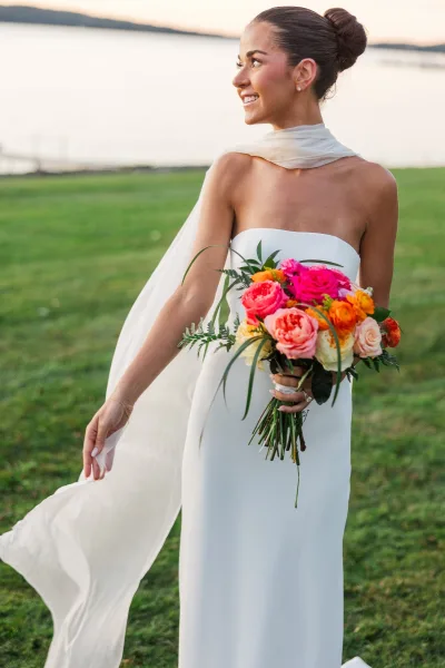 Bridal portrait of a bride holding bouquet, looking over her shoulder in a strapless dress and long veil by a lakeside at sunset