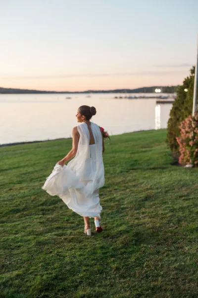 Bride portrait from behind walking away on grass, holding her dress and bouquet as a long veil blows by a lakeside at sunset