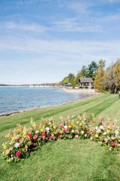 Outdoor ceremony decor with floral aisle decor in ground flower installations lining a grassy lakefront, sailboats and pavilion under blue sky