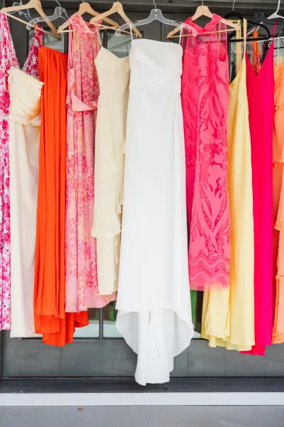 Bridesmaid dresses in mismatched bridesmaid dresses lineup on hangers, featuring floral, lace, and bright pink, orange, and yellow styles by window panes