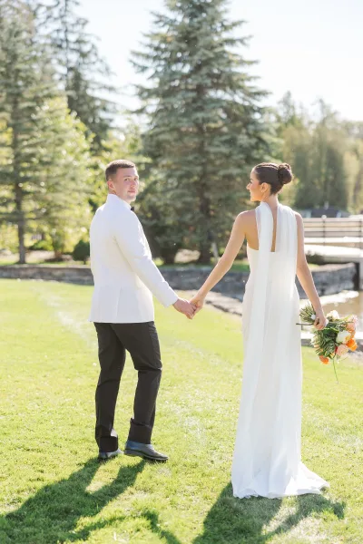 Couple portrait of bride and groom holding hands, walking away on a sunny lawn by a wooden bridge, bride with veil and bouquet