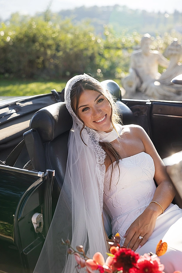 Bridal portrait of a bride in convertible holding a bright pink bouquet, veil draped over her shoulder with greenery and hills behind
