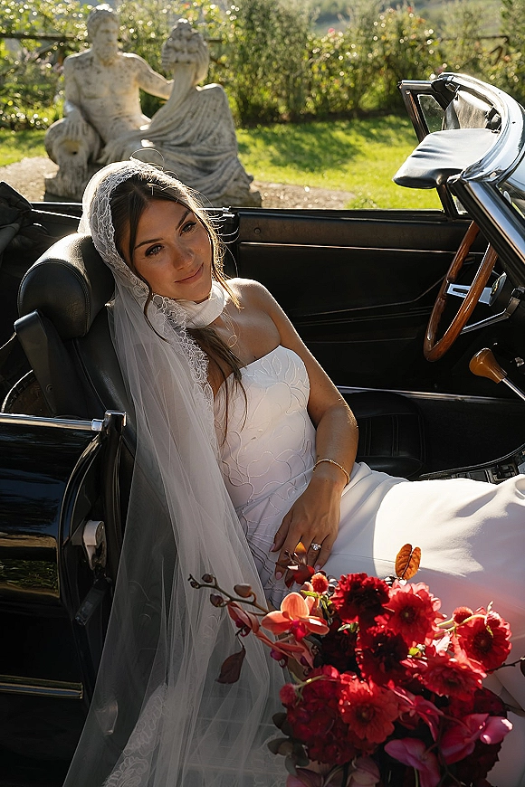 Bridal portrait of a bride in vintage car, wearing a strapless gown and lace veil, holding a red bouquet beside a sunlit garden statue