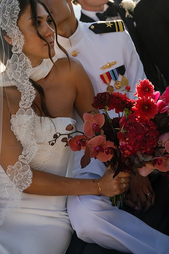 Couple portrait of bride in lace veil and strapless dress holding an orchid and red bouquet beside groom in military dress uniform with medals outdoors