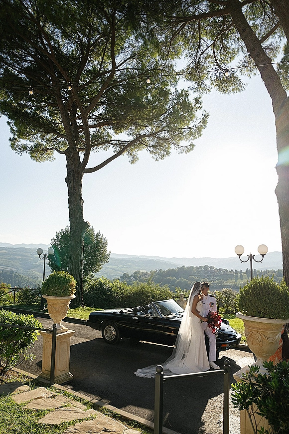 Couple portrait of bride in wedding dress and groom in military uniform kissing by a vintage convertible, mountains in the distance