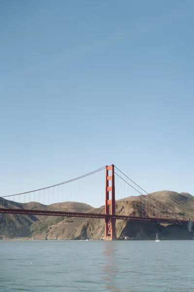 Suspension bridge, a red steel bridge, arches over blue water as a lone sailboat passes below, framed by coastal hills under a clear sky.
