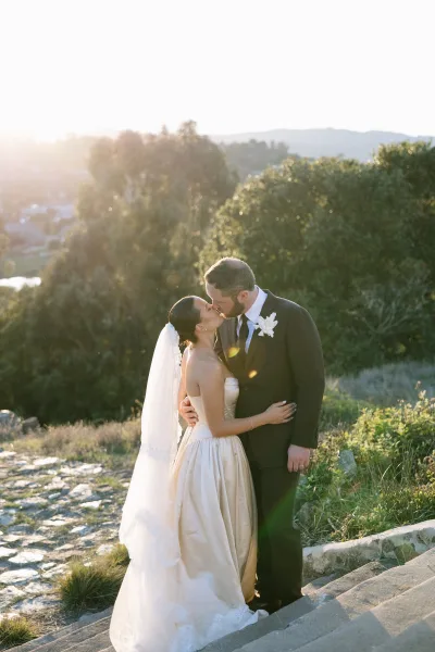 Wedding kiss portrait of a golden hour couple on stone steps, her lace-trim veil catching sunlight as trees line the hillside behind.