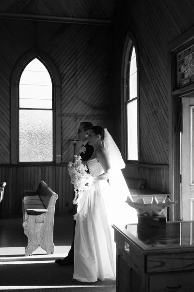 Wedding processional at a church ceremony entrance, bride entering with a cascading bouquet along the aisle framed by arched windows and wooden pews.