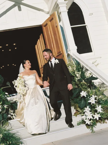 Ceremony exit with bride and groom walking down church steps, hand in hand, her bridal bouquet framed by wooden doors and white siding.
