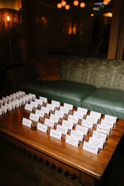 Escort card display of tented escort cards in calligraphy on a wood coffee table, with a sofa and pendant lights softly behind.