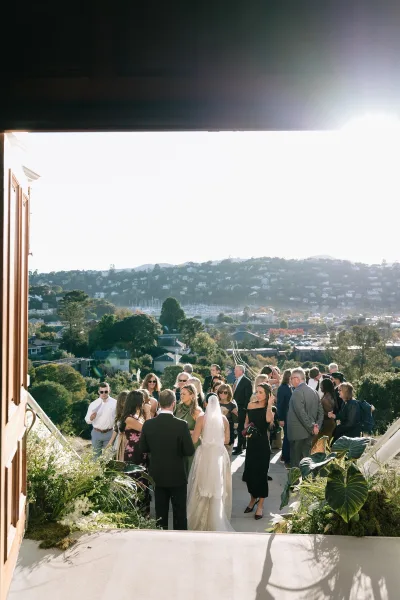 Wedding cocktail hour with guests mingling outdoors as the bride in a long bridal veil greets friends by a hillside view near the doorway