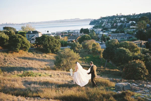 Couple portrait of a bride and groom walking on a grassy hillside overlooking a coastal town and water, her veil trailing.
