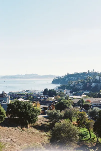Coastal town view over a harbor with sailboats, rooftops stepping down to a calm bay and coastline, with hills and distant mountains under clear sky.