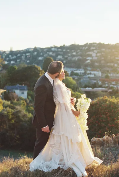 Couple portrait of the groom kissing the bride, her bridal veil catching light on a hillside with trees and town buildings under open sky.
