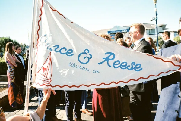 Wedding signage as a custom pennant flag featuring a sailboat graphic, displayed in an outdoor plaza with buildings and wedding guests.