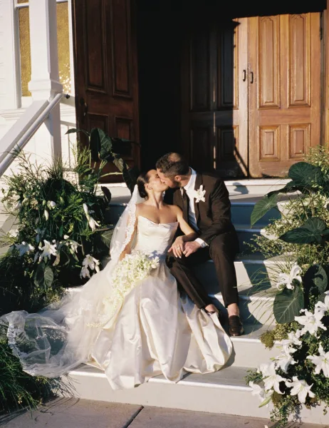 Wedding kiss portrait of a bride and groom on steps, her cascading bouquet between them, framed by wooden doors and a white exterior wall.