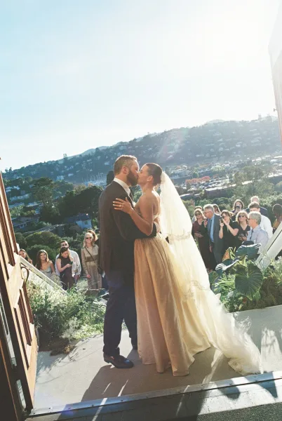 Wedding kiss portrait capturing a recessional kiss beneath a flowing bridal veil, backlit by sunlight at a wooden door as guests look on.