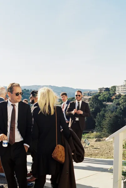 Wedding guests mingling on a sunny patio in sunglasses, chatting by a railing with hills and trees under a clear blue sky.