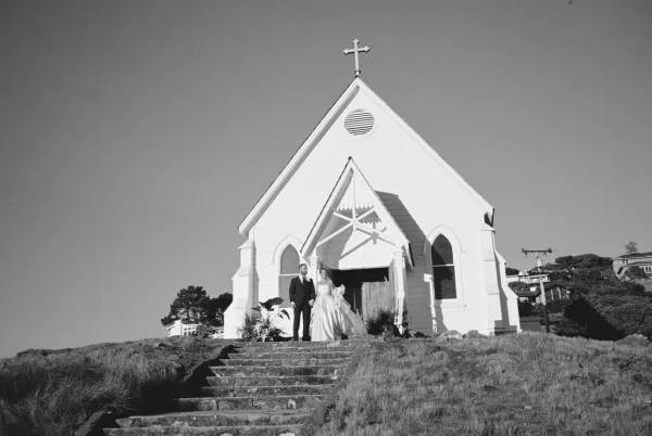 Couple portrait, a church wedding portrait on the stone steps of a chapel with arched windows, bride holding a bouquet beside a grassy hill.